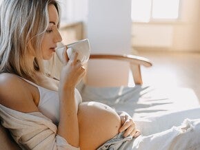 Фотографія вагітної жінки на дивані, що п'є з чашки й торкається живота