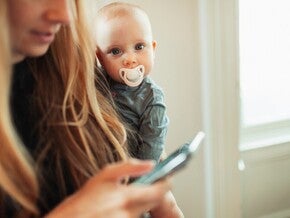 Жінка користується смартфоном, поки дитина з соскою виглядає їй через плече