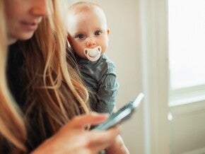 Фото: жінка користується смартфоном, поки дитина з соскою виглядає їй через плече